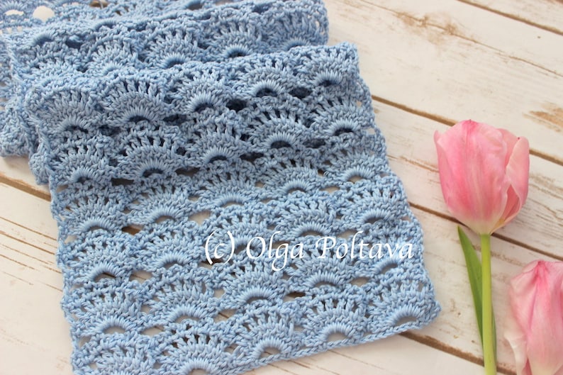 May include: A light blue crocheted scarf with a delicate, lacy pattern. The scarf is laid out on a white wooden surface with two pink tulips.