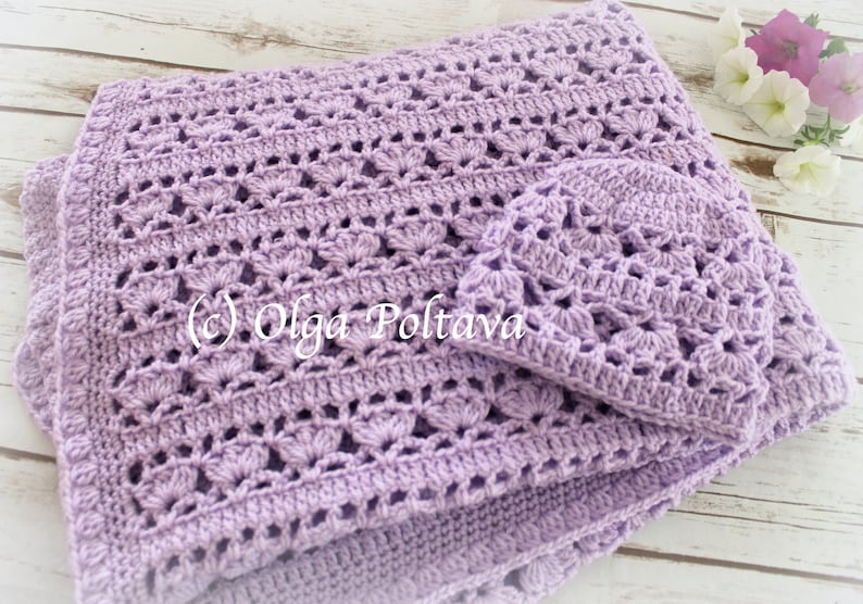 May include: A lavender crochet baby blanket and matching hat. The blanket has a delicate, lacy pattern and is folded in half. The hat is small and sits on top of the blanket.