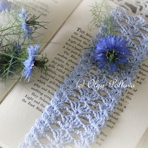 May include: A blue crocheted lace bookmark with a delicate floral pattern, resting on an open book with blue flowers. The text on the book reads "THE Blue the Bowery between the t Light does bric-a-brae, it for pain-ki The Blue modern pharm colates its on day pills are r pills rolled ou patula, rolle calcined magu board pill-bos which coveys play and beco soothing syrup Ikey Schoen Light and the East Side, "
