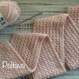 May include: A light pink crocheted scarf is displayed, alongside a ball of Premier Basix DK yarn and a crochet hook. The scarf is folded in a zig-zag pattern, showcasing its textured stitch. The yarn label is visible.