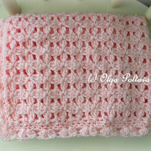 May include: A pink crocheted baby blanket with a delicate floral pattern. The blanket has a scalloped edge and is made with soft yarn.