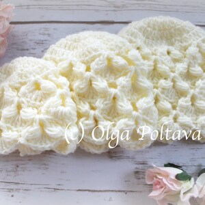 May include: Three crocheted hats in a cream color with a textured, raised pattern. The hats are arranged in a row on a white wooden surface.