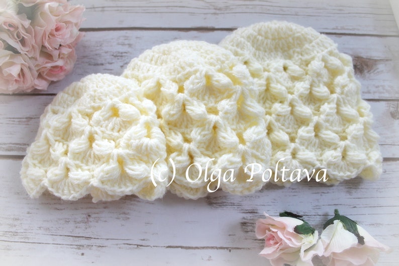 May include: Three crocheted hats in a cream color with a textured, raised pattern. The hats are arranged in a row on a white wooden surface.