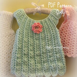 May include: A mint green crochet baby dress with a scalloped hem and a pink flower applique. The dress is hanging on a white hanger with a white ribbon. The text "PDF Pattern Only" and "Olga's Crochet Frenzy" are visible.