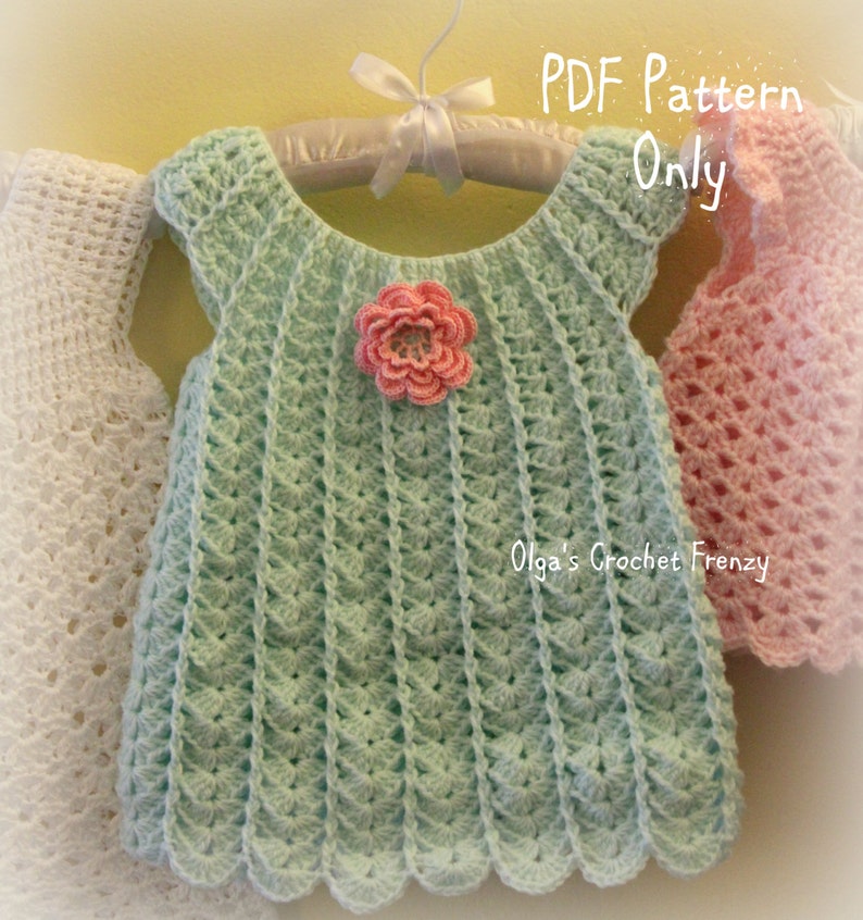 May include: A mint green crochet baby dress with a scalloped hem and a pink flower applique. The dress is hanging on a white hanger with a white ribbon. The text "PDF Pattern Only" and "Olga's Crochet Frenzy" are visible.