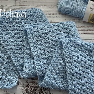 May include: A light blue crocheted scarf featuring an openwork stitch pattern. Beside the scarf lies a skein of light blue yarn with a label reading Soft & Shiny and a metal crochet hook. The scarf is folded in a series of three distinct sections.
