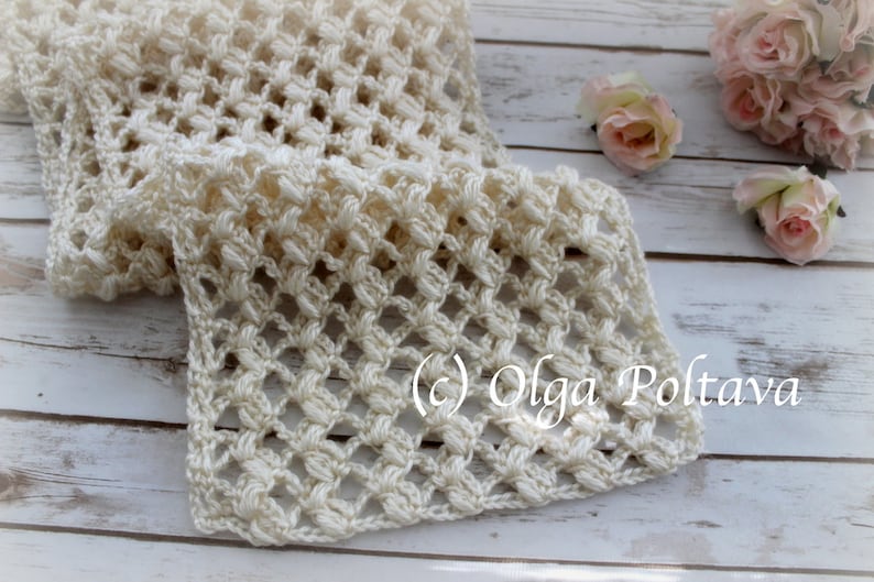 May include: A cream-coloured, open-weave crochet scarf. The scarf has a repeating pattern of raised stitches, creating a textured surface. The text "(c) Olga Poltava" is visible. The scarf is displayed on a light wooden surface.