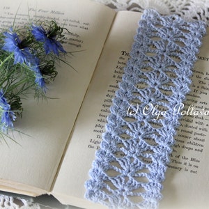 Op de afbeelding: Een lichtblauwe gehaakte bladwijzer met een delicaat opengewerkt patroon. De bladwijzer ligt op een open boek met een boeket blauwe bloemen op de linkerpagina.