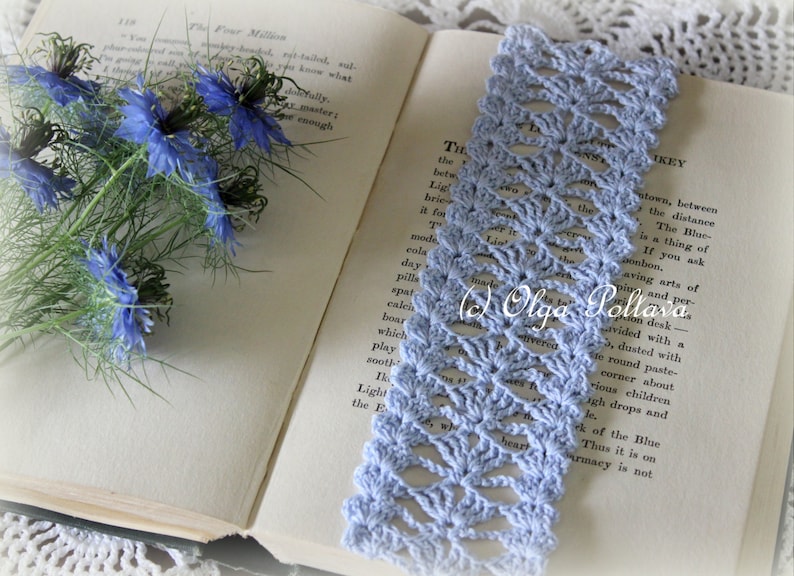 Op de afbeelding: Een lichtblauwe gehaakte bladwijzer met een delicaat opengewerkt patroon. De bladwijzer ligt op een open boek met een boeket blauwe bloemen op de linkerpagina.