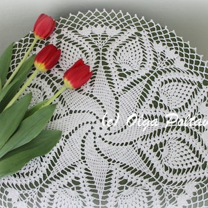 May include: A white, crocheted doily with a detailed, openwork design. Three red tulips with green stems and leaves are arranged on the doily. The doily has a circular shape and is placed on a green surface.