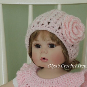 May include: A pink crocheted hat with a flower on the side. The hat is worn by a doll with brown hair and blue eyes. The doll is wearing a pink crocheted dress.