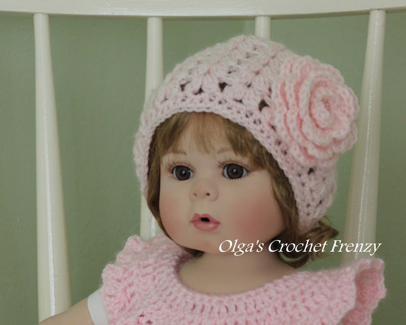 May include: A pink crocheted hat with a flower on the side. The hat is worn by a doll with brown hair and blue eyes. The doll is wearing a pink crocheted dress.