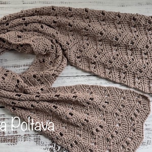 May include: A taupe-colored, open-weave crochet scarf. The scarf is made of soft yarn and features a repeating geometric pattern with small openings. The scarf is displayed on a light-colored wooden surface. The text "(c) Olga Poltava" is visible.