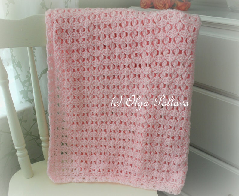 May include: A pink crocheted baby blanket with a delicate openwork pattern. The blanket is draped over a white chair.