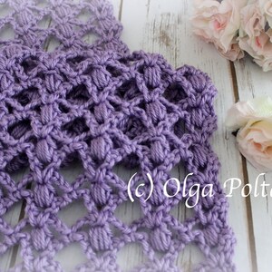 May include: A close-up of a crocheted lavender scarf with a textured, openwork design. The scarf features a repeating pattern of raised, rounded stitches. The background includes white flowers and a wooden surface. The text "(c) Olga Poltava" is visible.