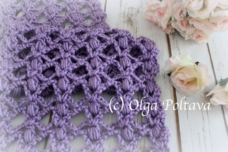 May include: A close-up of a crocheted lavender scarf with a textured, openwork design. The scarf features a repeating pattern of raised, rounded stitches. The background includes white flowers and a wooden surface. The text "(c) Olga Poltava" is visible.
