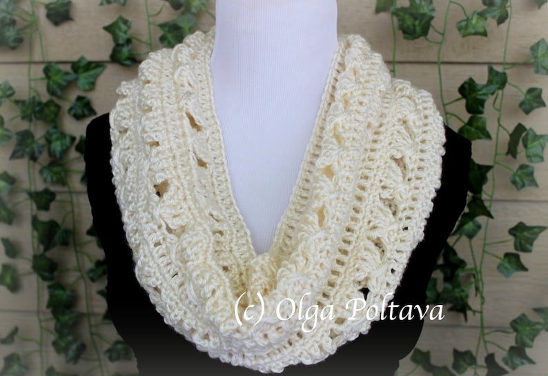 May include: An off-white crochet infinity scarf featuring a textured, repeating pattern of raised stitches and openwork loops. The scarf is draped around a white mannequin neck form, displaying the intricate yarn work and cream color.