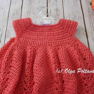 May include: A coral-colored, handmade crochet dress for a baby or toddler. The dress is displayed on a white hanger against a light wood background. The dress features a detailed stitch pattern and a round neckline. The copyright notice "(c) Olga Poltava" is visible.
