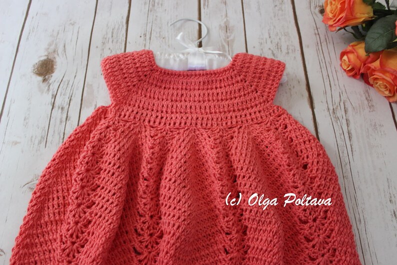 May include: A coral-colored, handmade crochet dress for a baby or toddler. The dress is displayed on a white hanger against a light wood background. The dress features a detailed stitch pattern and a round neckline. The copyright notice "(c) Olga Poltava" is visible.