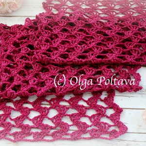 May include: A handmade, open-weave scarf in a deep magenta color. The scarf is crocheted with a delicate, lacy pattern, and the texture is visible. The scarf is displayed on a light-colored wooden surface, with floral accents.