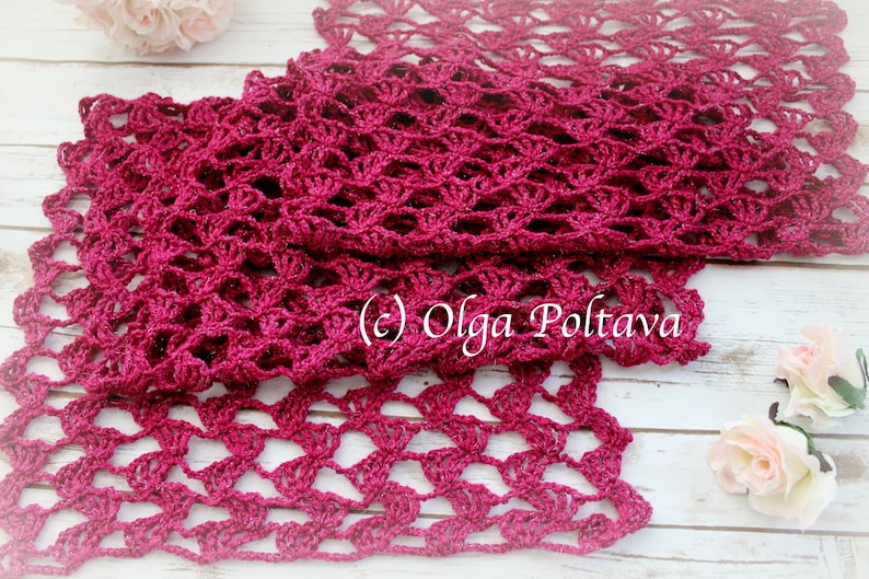 May include: A handmade, open-weave scarf in a deep magenta color. The scarf is crocheted with a delicate, lacy pattern, and the texture is visible. The scarf is displayed on a light-colored wooden surface, with floral accents.
