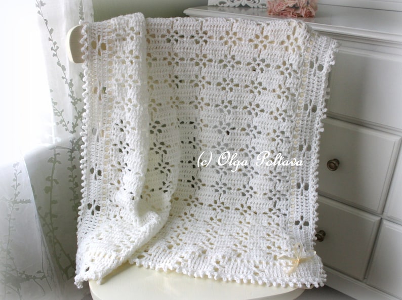 May include: White crocheted baby blanket with a delicate floral pattern. The blanket is draped over a white chair and has a small white bow on the corner.