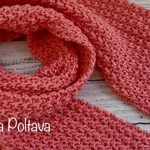 Può includere: Una sciarpa lavorata a maglia color corallo, con un motivo a punto strutturato, è mostrata in primo piano. La sciarpa è drappeggiata e piegata, mostrando la sua lunghezza e la lavorazione a maglia dettagliata. Lo sfondo è una superficie di legno di colore chiaro.