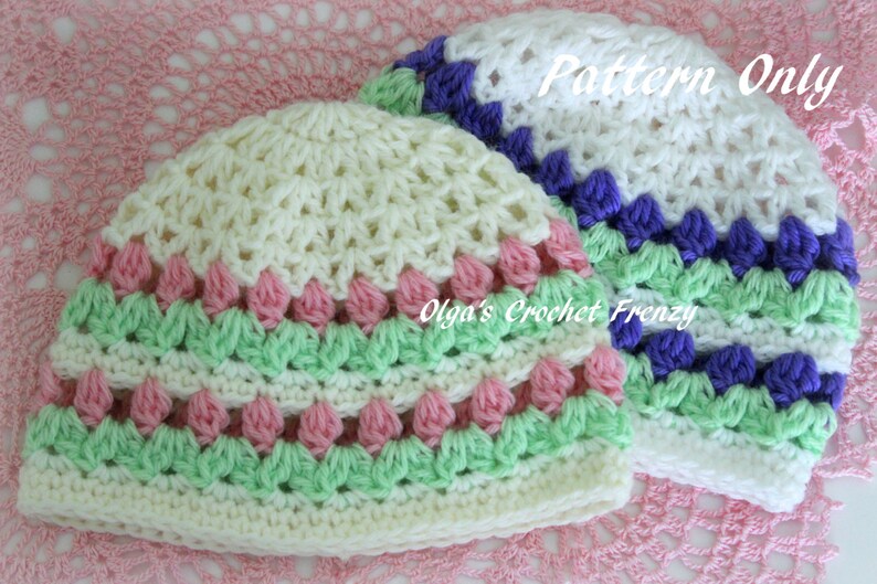 Puede incluir: Dos gorros de ganchillo. Uno es de color crema con detalles en rosa y verde. El otro es blanco con detalles en morado y verde. El texto "Pattern Only" es visible en el gorro blanco. Los gorros est&aacute;n hechos a mano.