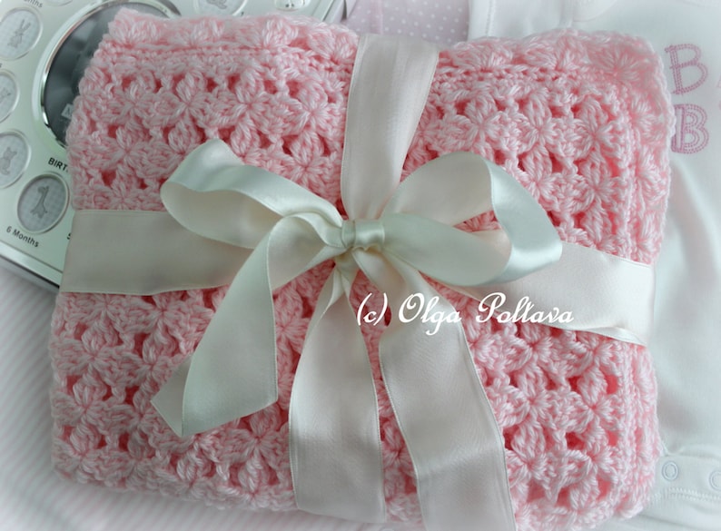 May include: A pink crocheted baby blanket with a white satin ribbon tied in a bow. The blanket has a delicate floral pattern.
