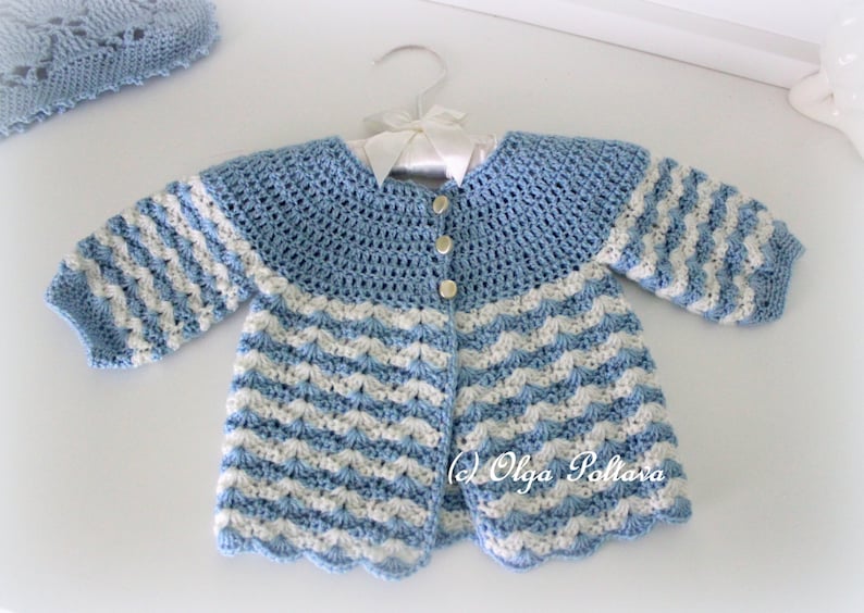 May include: A handmade baby cardigan in light blue and white crochet. The cardigan features a scalloped hem and sleeves with alternating stripes. The collar is a solid light blue, and the cardigan fastens with three buttons. A matching bonnet is visible in the background.