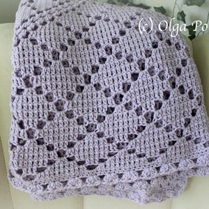 Puede incluir: Una manta de ganchillo de color lavanda con un delicado patr&oacute;n de encaje. La manta est&aacute; doblada y drapeada sobre un sill&oacute;n blanco.