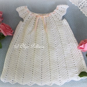 May include: White crocheted baby dress with a pink ribbon at the neckline. The dress has a scalloped hem and a delicate lace pattern.