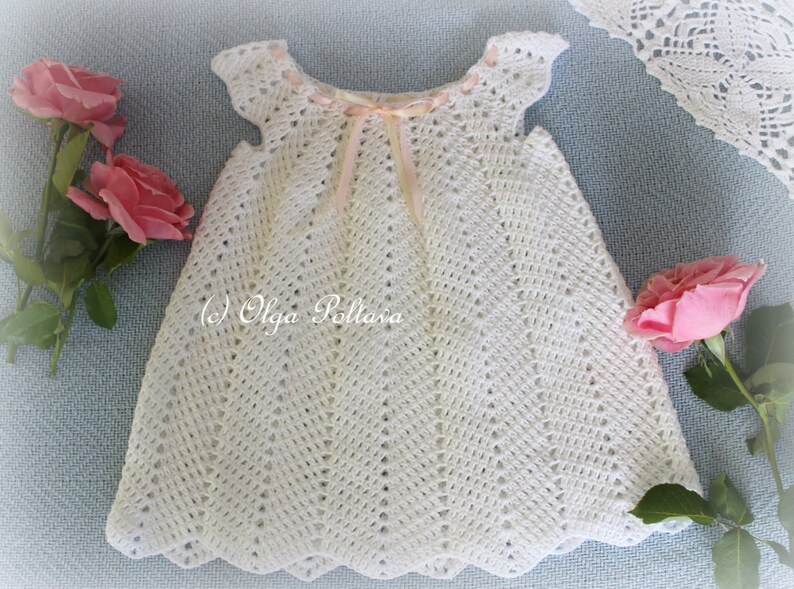 May include: White crocheted baby dress with a pink ribbon at the neckline. The dress has a scalloped hem and a delicate lace pattern.