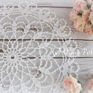 May include: A white, handmade crochet doily with a detailed floral pattern. The doily is circular and sits on a light-coloured wooden surface, with a bouquet of pink roses in the background. The text "(c) Olga Poltava" is visible.
