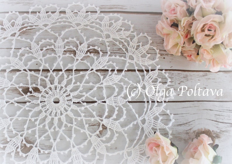 May include: A white, handmade crochet doily with a detailed floral pattern. The doily is circular and sits on a light-coloured wooden surface, with a bouquet of pink roses in the background. The text "(c) Olga Poltava" is visible.