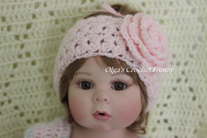 May include: A pink crocheted headband with a large pink crocheted flower on top. The headband is worn by a doll with brown hair and blue eyes.