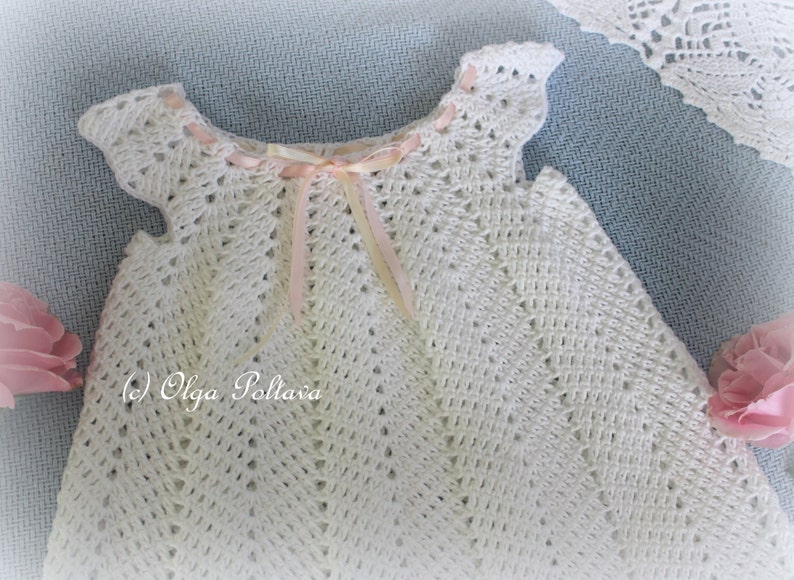 May include: White crocheted baby dress with a pink ribbon tied around the neckline. The dress has a delicate, lacy pattern and is perfect for a special occasion.