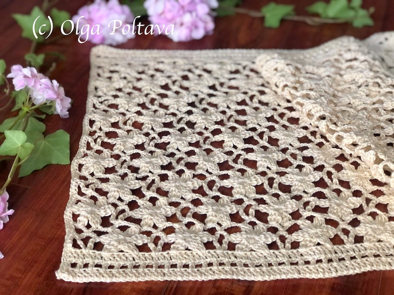 May include: A crocheted table runner with a delicate floral pattern in a cream color. The runner is laid out on a wooden table.