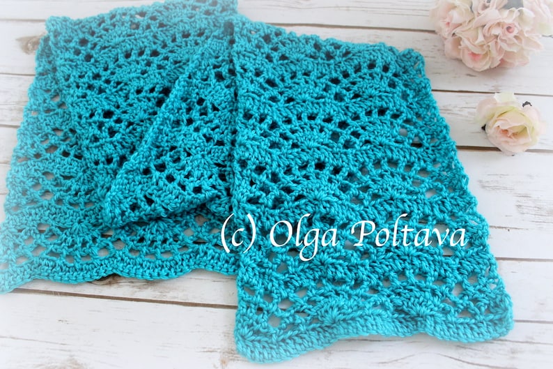 May include: A teal blue crocheted scarf with a lacy pattern. The scarf is folded in half and laid on a white wooden surface.