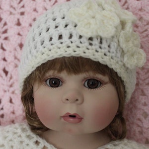 Op de afbeelding: Pop met een witte gehaakte hoed met een bloemaccent en een bijpassende gehaakte top. De pop heeft bruine ogen, roze wangen en lichtbruin haar. De tekst "Olga's Crochet Frenzy" is zichtbaar.