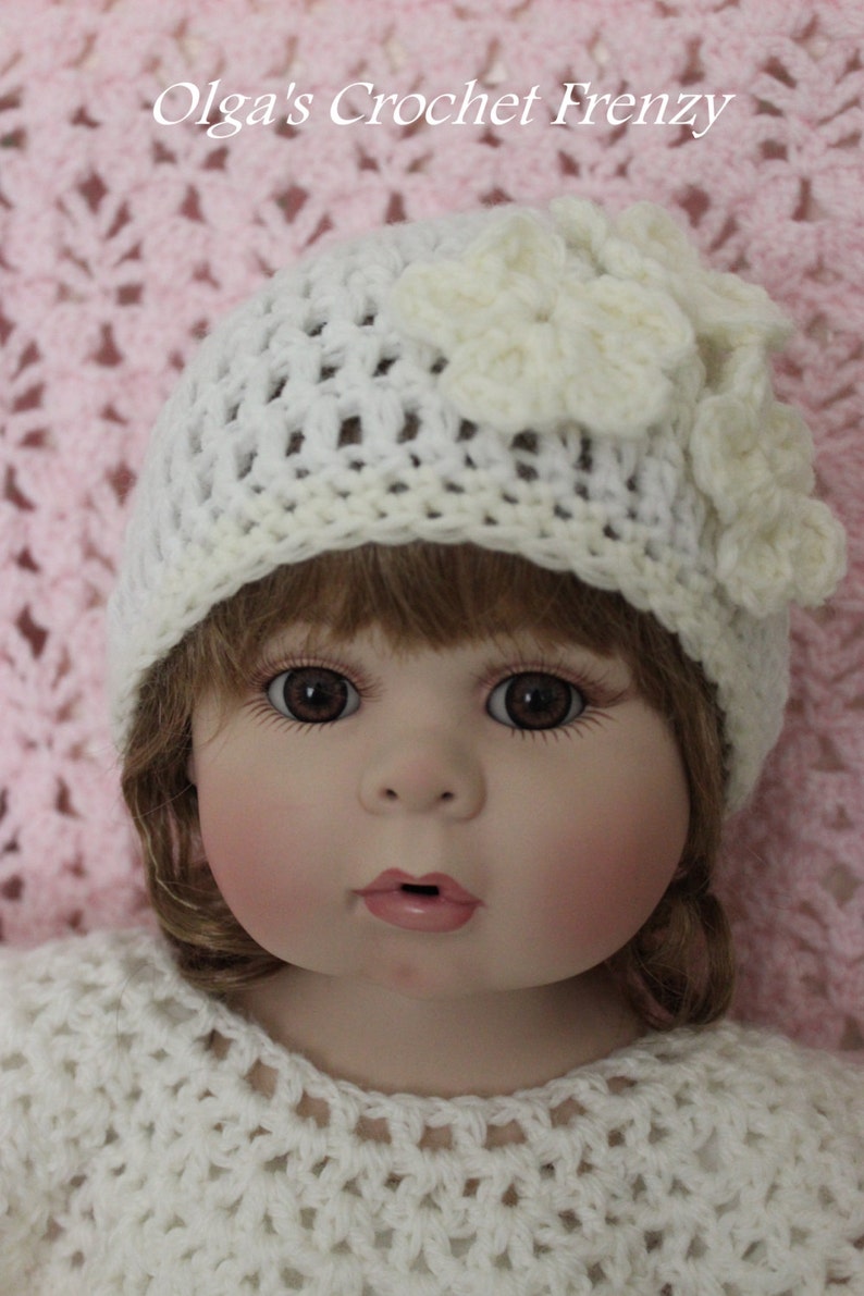 Op de afbeelding: Pop met een witte gehaakte hoed met een bloemaccent en een bijpassende gehaakte top. De pop heeft bruine ogen, roze wangen en lichtbruin haar. De tekst "Olga's Crochet Frenzy" is zichtbaar.