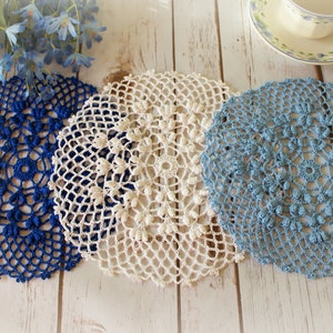 May include: Three circular crochet doilies in dark blue, cream, and light blue. Each doily features an intricate, openwork lace pattern with a central ring and radiating floral-like motifs, crafted with fine thread in a delicate, decorative style.