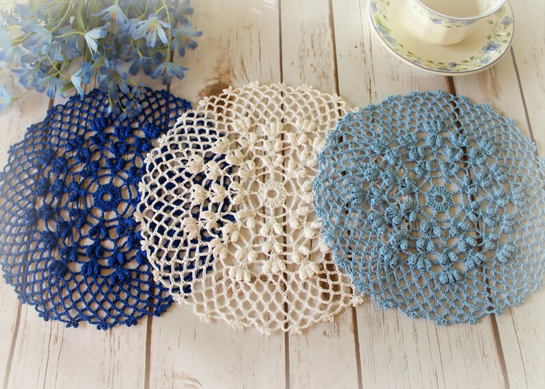 May include: Three circular crochet doilies in dark blue, cream, and light blue. Each doily features an intricate, openwork lace pattern with a central ring and radiating floral-like motifs, crafted with fine thread in a delicate, decorative style.