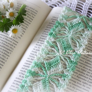 May include: A green and white crocheted bookmark with a delicate floral design. The bookmark is lying open on a book with text. The bookmark has the text "Olga Poltava" in the lower right corner.