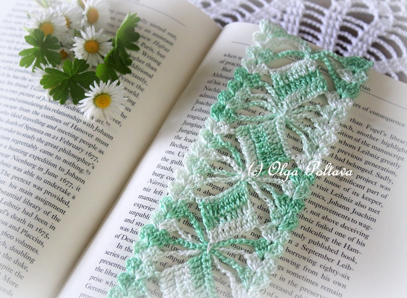 May include: A green and white crocheted bookmark with a delicate floral design. The bookmark is lying open on a book with text. The bookmark has the text "Olga Poltava" in the lower right corner.