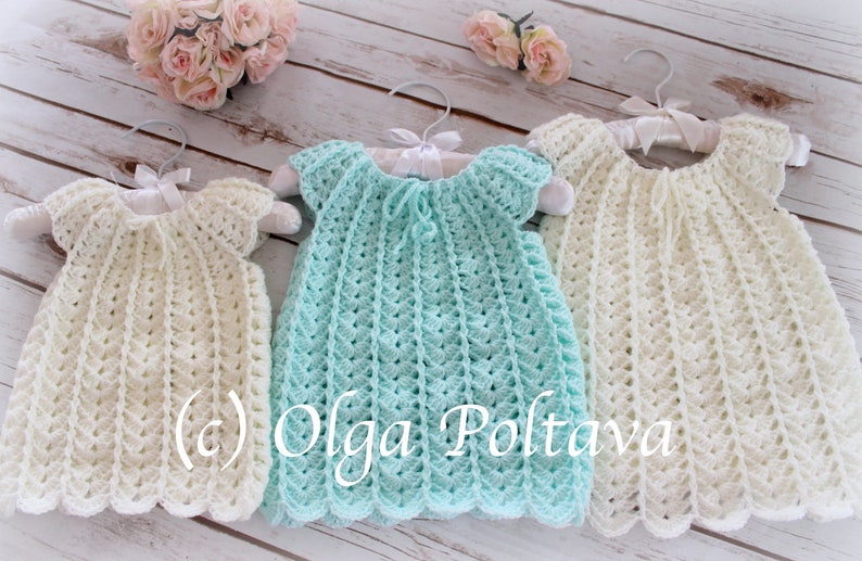 May include: Three handmade crochet dresses in cream and light blue hang on white hangers. The dresses have a scalloped hem and a textured stitch pattern. The dresses are displayed on a light wood surface with a floral arrangement in the background.