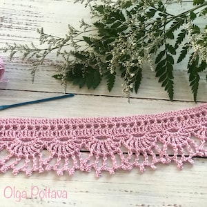 Puede incluir: Primer plano de un ribete de encaje de ganchillo rosa con un borde festoneado. El ribete está sobre una superficie de madera blanca con un ovillo de hilo rosa y un ganchillo.