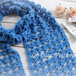 Pode incluir: Uma echarpe de croch&ecirc; azul com um padr&atilde;o rendado. A echarpe est&aacute; dobrada e colocada sobre uma superf&iacute;cie de madeira branca. O texto "Knit to Cozie" &eacute; vis&iacute;vel em uma etiqueta branca.