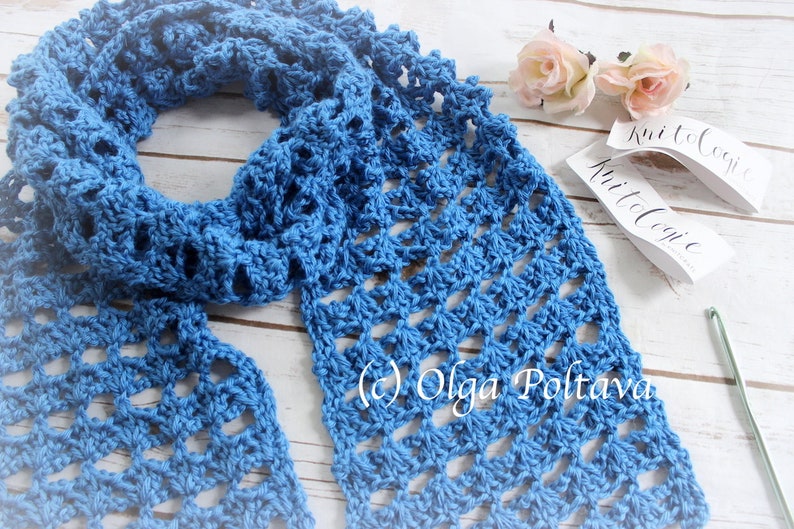 Pode incluir: Uma echarpe de croch&ecirc; azul com um padr&atilde;o rendado. A echarpe est&aacute; dobrada e colocada sobre uma superf&iacute;cie de madeira branca. O texto "Knit to Cozie" &eacute; vis&iacute;vel em uma etiqueta branca.