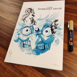 Peut inclure: Une impression d'art rectangulaire blanche avec des illustrations à l'encre bleue de personnages de Spirited Away, dont un grand visage, un dragon et des silhouettes. Le texte Spirited Away est imprimé en haut. Un marqueur doré est posé à côté.